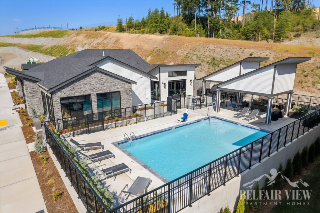 Modern apartment complex with a rectangular blue swimming pool, surrounded by a black metal fence and white concrete deck with lounge chairs and umbrellas.