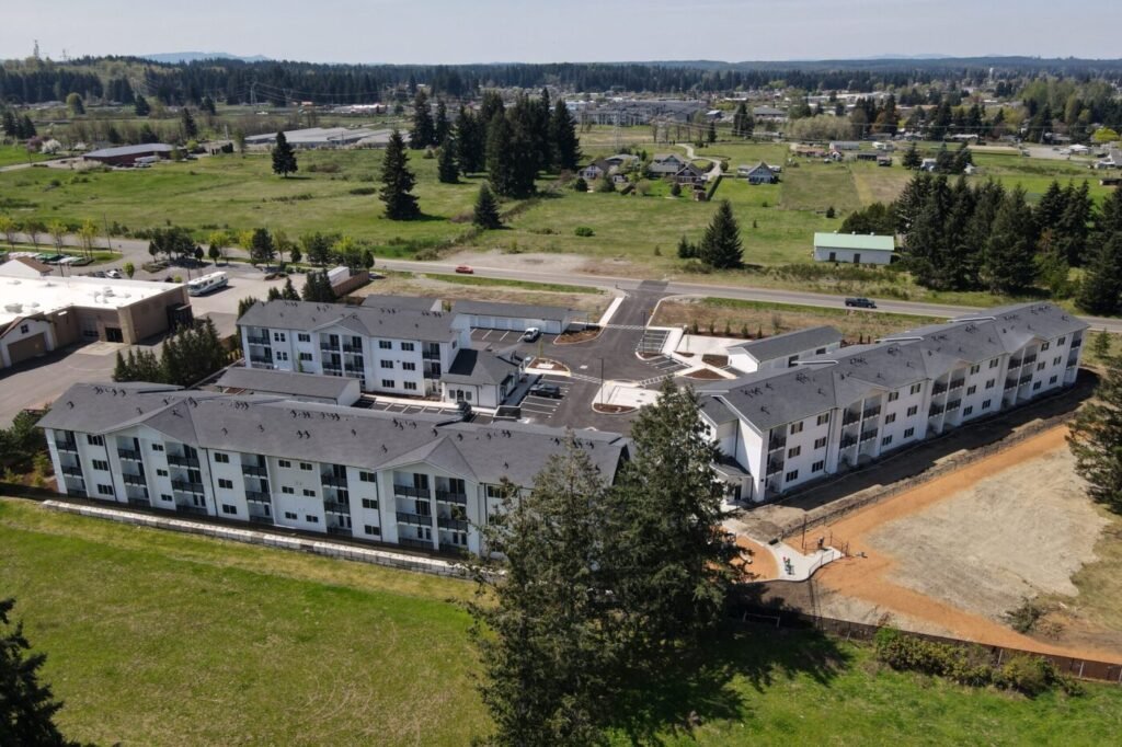 Aerial view of a U-shaped apartment complex with white buildings and gray roofs, surrounded by green fields.