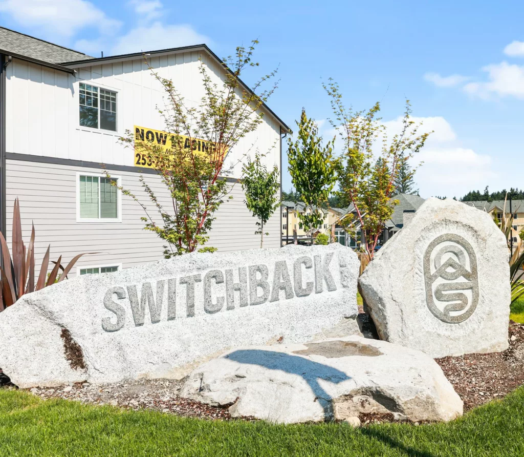Stone sign reading SWITCHBACK with a circular logo in front of a residential complex and young trees nearby, under a blue sky.