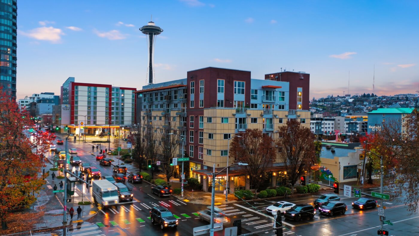 Evening street scene in downtown Seattle with the Space Needle visible in the distance and a wet, busy intersection below.