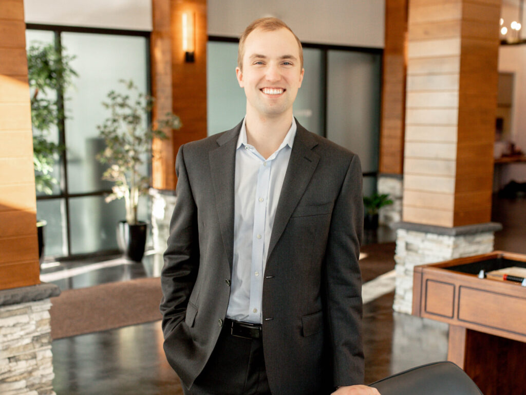 Smiling man in a dark suit and light blue shirt standing in a modern office lobby with wood panels and plants.