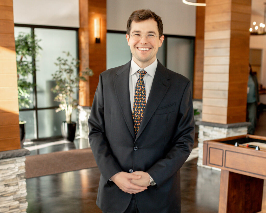 Smiling man in a dark suit with a patterned tie stands in a modern lobby with wood panels and plants in the background, hands clasped in front.