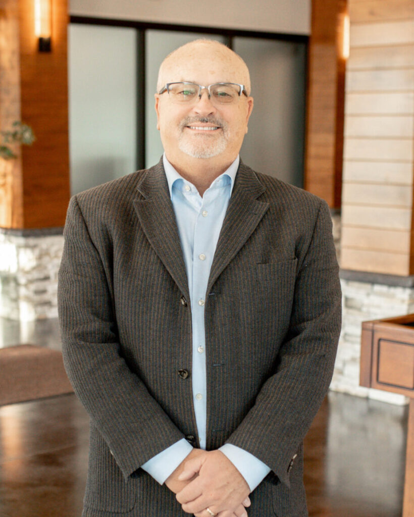 Smiling man in a dark pinstripe blazer and light blue shirt standing in a modern lobby.