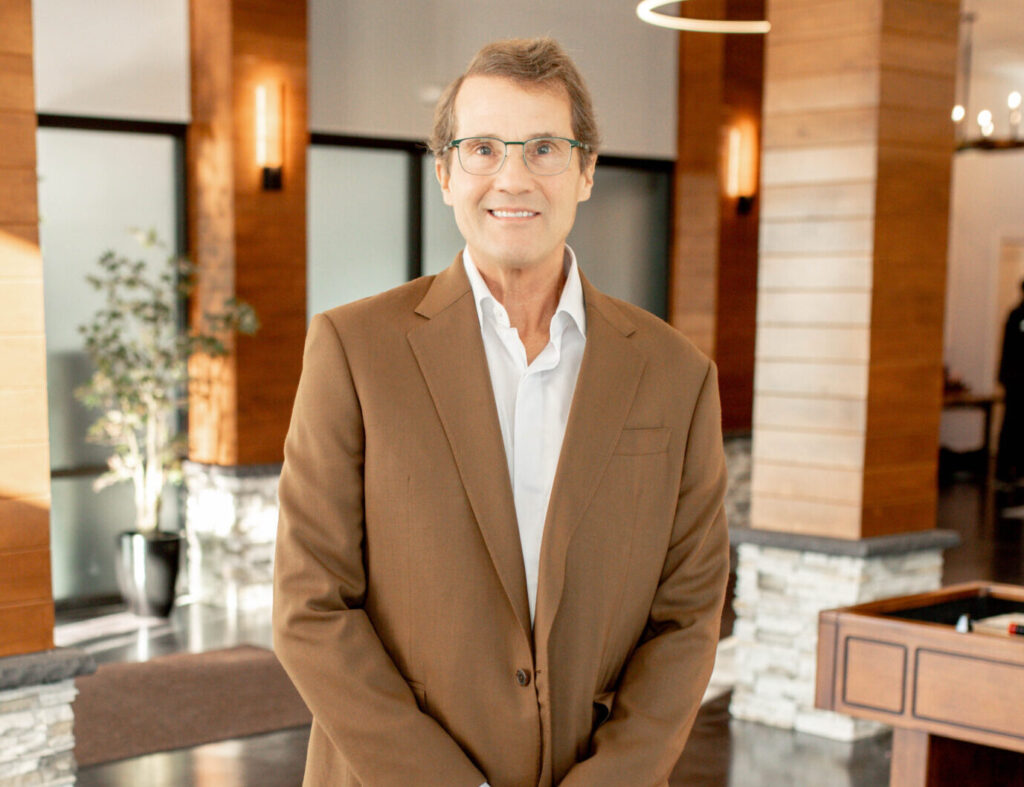 A smiling man in a brown suit and white shirt posing in a modern lobby with wood panels and stone accents.